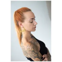 Donna con orecchini Blessed Hoops dorati e tatuaggi di profilo su sfondo chiaro
