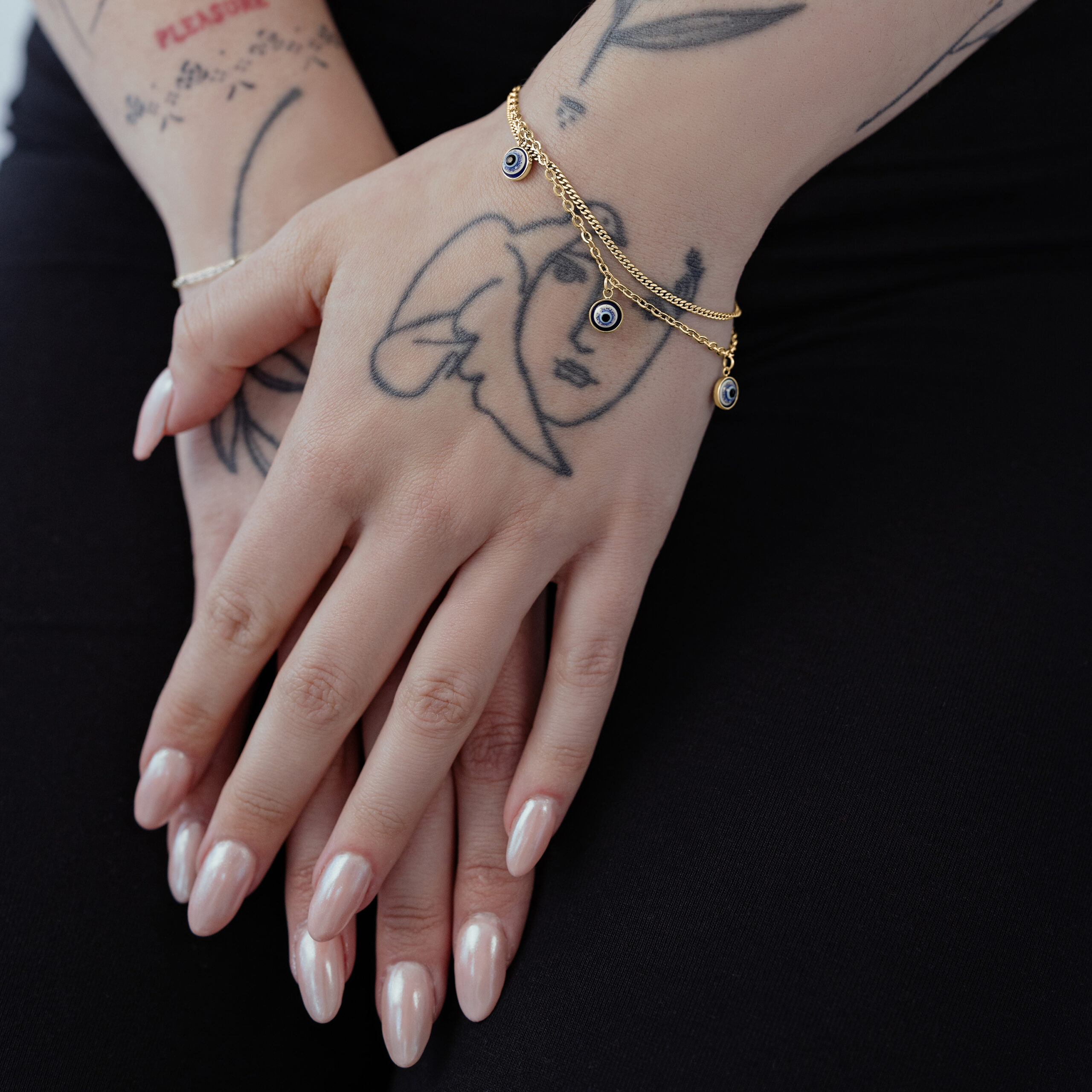 Little Nazar Armband in Gold auf Hand Goldenes Little Nazar Armband um ein Handgelenk mit Tattoos auf schwarzem Hintergrund